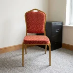 Fabric stackable conference chair featuring patterned red and gold patterned fabric upholstery, high-density padded seat and crown-back cushion, an armless space-saving stackable frame, an integrated top hand-hold, and a gold powder-coated square steel tube frame with bumper guards and floor glides.