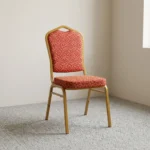 Fabric stackable conference chair featuring patterned red and gold patterned fabric upholstery, high-density padded seat and crown-back cushion, an armless space-saving stackable frame, an integrated top hand-hold, and a gold powder-coated square steel tube frame with bumper guards and floor glides.