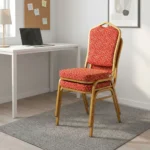 Fabric stackable conference chair featuring patterned red and gold patterned fabric upholstery, high-density padded seat and crown-back cushion, an armless space-saving stackable frame, an integrated top hand-hold, and a gold powder-coated square steel tube frame with bumper guards and floor glides.