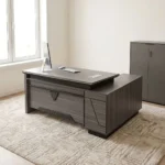 Executive 2.0m L-shaped office desk featuring a dark grey wood-grain laminate finish, dual flip-top desktop cable management grommets, a stylized front modesty panel with black geometric accent detailing, a integrated side return extension unit with drawer storage, and solid panel-style engineered wood base supports.