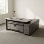 Executive 2.0m L-shaped office desk featuring a dark grey wood-grain laminate finish, dual flip-top desktop cable management grommets, a stylized front modesty panel with black geometric accent detailing, a integrated side return extension unit with drawer storage, and solid panel-style engineered wood base supports.