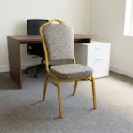 Comfortable armless fabric conference chair featuring patterned beige fabric upholstery over a thick padded seat cushion and backrest, an armless stackable crown-back frame design with an integrated top hand-hold handle, and a durable gold powder-coated square steel four-legged base.