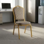 Comfortable armless fabric conference chair featuring patterned beige fabric upholstery over a thick padded seat cushion and backrest, an armless stackable crown-back frame design with an integrated top hand-hold handle, and a durable gold powder-coated square steel four-legged base.