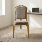 Fabric conference event chair featuring patterned beige fabric upholstery over a thick padded seat cushion and backrest, an armless stackable crown-back frame design with an integrated top hand-hold handle, and a durable gold powder-coated square steel four-legged base.