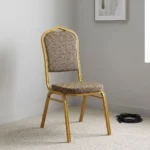 Fabric conference event chair featuring patterned beige fabric upholstery over a thick padded seat cushion and backrest, an armless stackable crown-back frame design with an integrated top hand-hold handle, and a durable gold powder-coated square steel four-legged base.