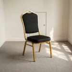 Black stackable event chair featuring black patterned fabric upholstery with gold dot accents on a crown-back backrest and padded seat cushion, an armless space-saving frame, and a durable gold powder-coated aluminum steel tubular frame base with stack bumpers and floor glides.