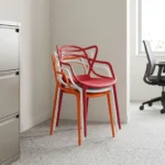 Outdoor stackable plastic chair featuring a weather-resistant molded polypropylene plastic body with an intricate interlocking loop backrest and integrated armrests, a contoured seat pan, and a space-saving stackable structure available in orange, white, and red finishes, supported by four integrated tapered legs with non-marking floor glides.