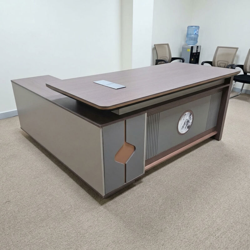 1.8m executive office table featuring a walnut wood grain laminate desktop with bevel-trimmed edges and grey side paneling, an L-shaped return cabinet structure with integrated storage compartments, a front modesty panel with fluted detailing and a central decorative emblem, built-in desktop flip-top cable management grommet, and a heavy-duty full-plinth base with floor-protective glides.