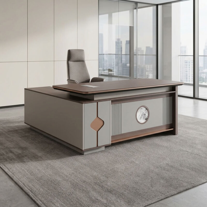 1.8m executive office table featuring a walnut wood grain laminate desktop with bevel-trimmed edges and grey side paneling, an L-shaped return cabinet structure with integrated storage compartments, a front modesty panel with fluted detailing and a central decorative emblem, built-in desktop flip-top cable management grommet, and a heavy-duty full-plinth base with floor-protective glides.