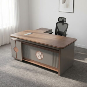 1.8m executive office table featuring a walnut wood grain laminate desktop with bevel-trimmed edges and grey side paneling, an L-shaped return cabinet structure with integrated storage compartments, a front modesty panel with fluted detailing and a central decorative emblem, built-in desktop flip-top cable management grommet, and a heavy-duty full-plinth base with floor-protective glides.